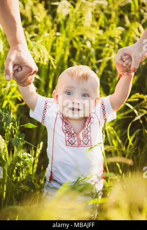 Portrait of ethnic ukrainian infant baby standing at sunny green rye background. Father, mother holding little hands of child. Vertical color photogra Stock Photo