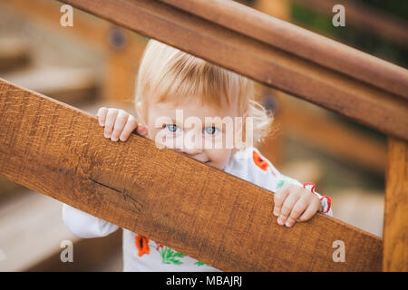 1 year old little caucasian girl palaing hide and seek game outdoors on sunny warm summer day. Closeup portrait of tricky face of blonde baby hiding f Stock Photo