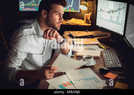 Young bearded man trader at office sitting at table looking at monitor concerned holding eyeglasses and document paper Stock Photo