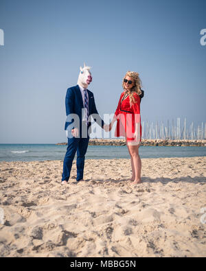 Unusual couple have marriage on the beach. Elegant man in funny horse mask and suit with cute young woman in bright red dress Stock Photo
