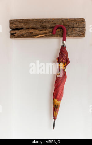 Red and yellow umbrella, closed, hung from a wooden coat rack on a white wall Stock Photo