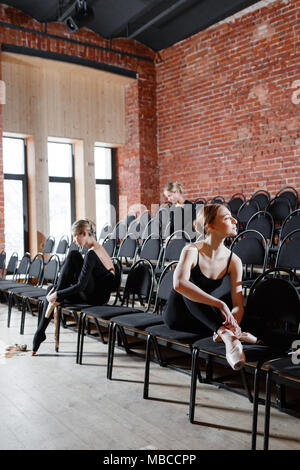 The ballet concept. Young ballerina girls sitting on the black chairs in the hall. Women at the rehearsal in black bodysuits. Prepare a theatrical performance Stock Photo