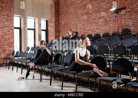 The ballet concept. Young ballerina girls sitting on the black chairs in the hall. Women at the rehearsal in black bodysuits. Prepare a theatrical performance Stock Photo