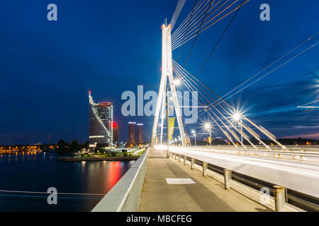 Riga, Latvia. Vansu Cable-Stayed Bridge In Bright Night Illumination Over The Daugava River, Skyscrapers Of Downtown And Blue Sky Background Stock Photo