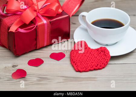 Red gift box with ribbon on a wooden background. A cup of coffee and a knitted heart. Valentine's Day. Stock Photo