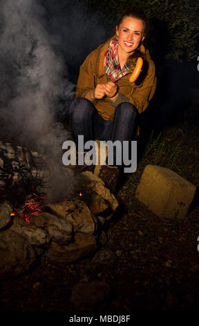 Into the wild. smiling traveller woman by a bonfire grilling sausage Stock Photo