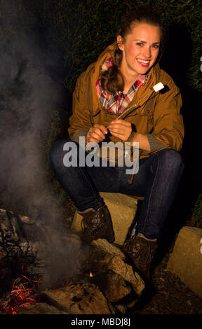 Into the wild. happy young traveller woman by the camping fire with roasted marshmallow Stock Photo