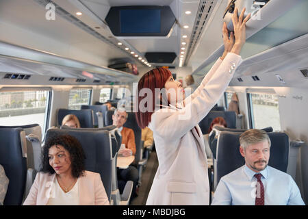 Businesswoman stowing suitcase in overhead compartment on passenger train Stock Photo