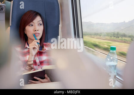Thoughtful young woman writing in journal and looking out window on passenger train
