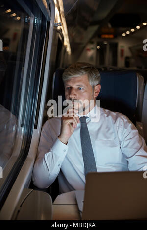 Serious, thoughtful businessman working at laptop, looking out window on passenger train at night Stock Photo