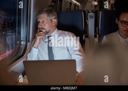Serious, thoughtful businessman working at laptop, looking out window on passenger train at night Stock Photo