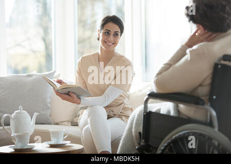 Young caregiver reading a book to a senior woman in a wheelchair Stock Photo