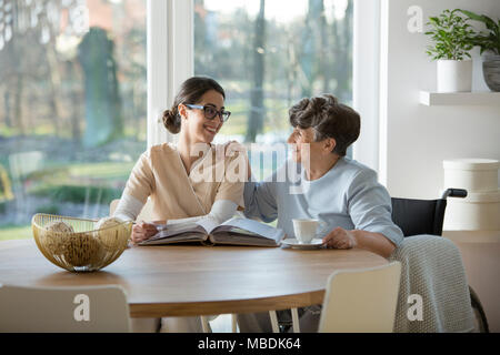 Happy pensioner and nurse sitting together at the table and having a conversation Stock Photo