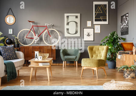 Red bike on a wooden cabinet, green armchairs and posters on the grey wall in a messy living room interior Stock Photo