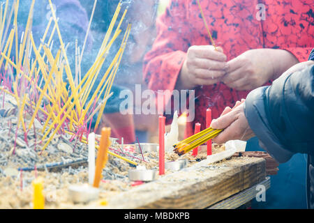 Close-up view of the hands of a woman wearing an asian style blouse lighting incense sticks before sticking them into sand during a buddhist ceremony. Stock Photo