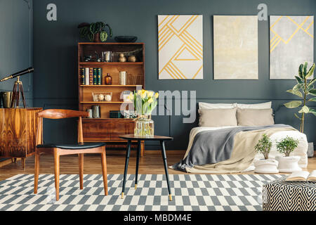 Bed, plants, white and grey carpet, pouf, chairs, vintage bookcase, telescope and grey wall in retro bedroom interior Stock Photo