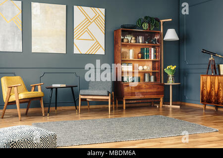 Bright living room interior with retro bookcase, chest of drawers, chairs, grey carpet, modern paintings on dark wall and pouf Stock Photo