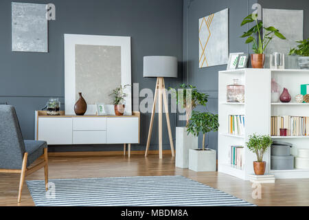 Plants next to lamp in grey living room interior with paintings and books on shelves Stock Photo
