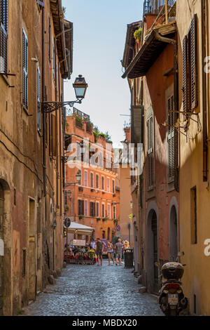An iconic Italian scene with tourists walking along a cobbled street in the Trastevere part of Rome, Italy, past a restaurant with people sat outside  Stock Photo