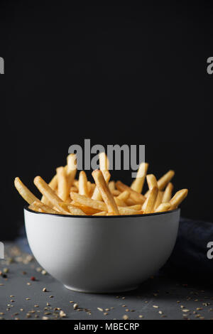 some appetizing french fries served in a white ceramic bowl, placed on a gray rustic wooden table, against a black background with some blank space on Stock Photo