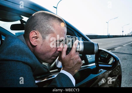 closeup of a young caucasian detective or paparazzi man, in a gray suit, taking photos from inside a car Stock Photo