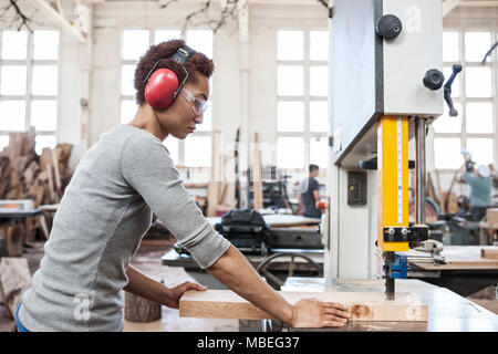 Black woman factory worker using a band saw to cut wood in a woodworking factory. Stock Photo