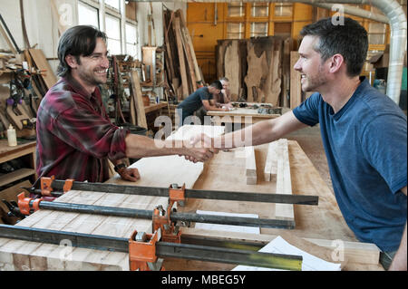 Team of four men, factory workers at a work station in a large woodworking factory, two shaking hands. Stock Photo