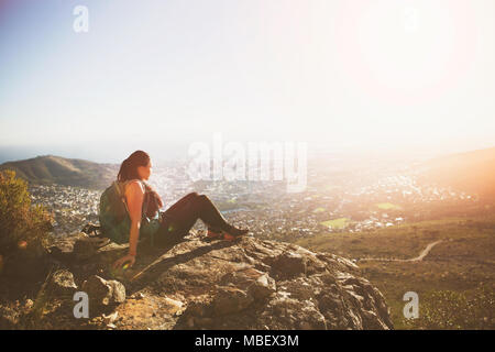 Serene female backpacker sitting on rocks above sunny city Stock Photo