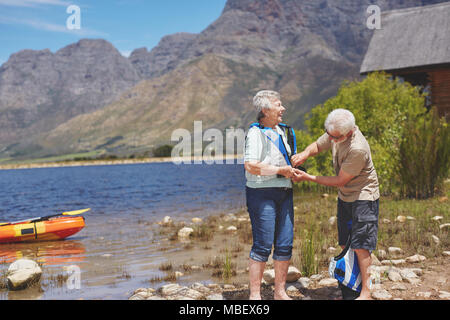 Active senior couple putting on life jackets, preparing for kayaking at sunny summer lakeside Stock Photo