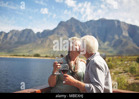 Romantic, active senior couple kissing and drinking wine on sunny summer balcony at lakeside Stock Photo
