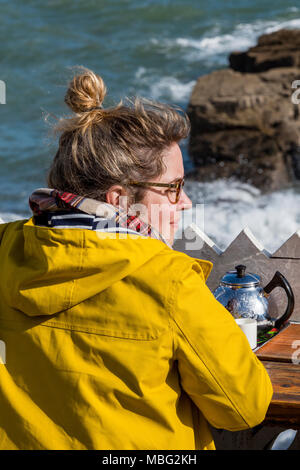 A woman or female sitting drinking tea with a yellow coast and teapot at the seaside on a rocky beach. Woman dressed in yellow fishermans jacket smock Stock Photo