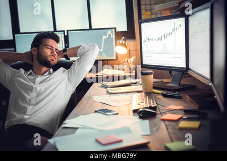 Serious trader leaning on chair holding hands behind head looking at stock market data graph on computer monitors relaxing in office feeling fatigued  Stock Photo