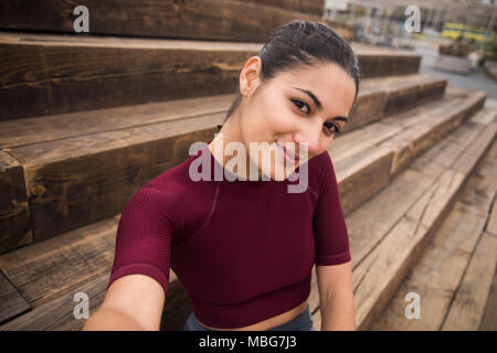 Charming young woman smiling at the camera wearing a maroon shirt and hair tied back while taking selfie Stock Photo