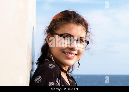 Portrait of an attractive and young woman (mixed ethnicities of Caucasian and Nepalese) wearing glasses and smiling at the ocean. Stock Photo