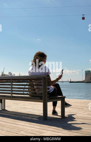 Portrait of a young woman sitting on a bench and looking at her smartphone in the port of Barcelona with the red cabin of a cableway in the background. Stock Photo