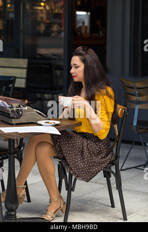 Young female with dark and long brown hair, wearing yellow shirt and brown skirt with white polka dots, sitting in a coffee shop, drinking from white  Stock Photo