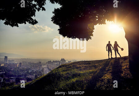 Yong couple in silhouette holding by hands and looking at the sunset city landscape Stock Photo
