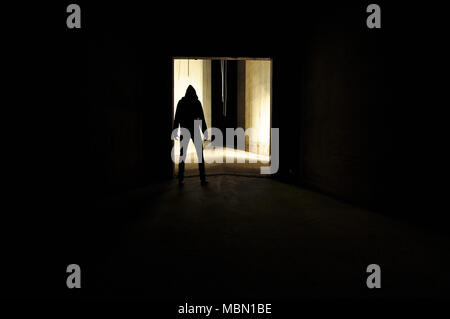 unrecognizable silhouette of person with a hood standing in a door way of an empty concrete room. Stock Photo