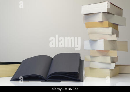 Black open book in foreground of pile of books on the white table and light grey background Stock Photo