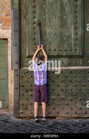 A young boy banging the door knocker on Porta del Popolo is a gate of the Aurelian Walls and the entrance to the North of the city in Rome, Italy. The Stock Photo