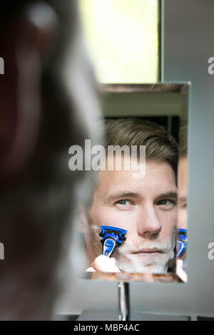 Reflection of handsome man with green eyes shaving with blue razor in small mirror Stock Photo