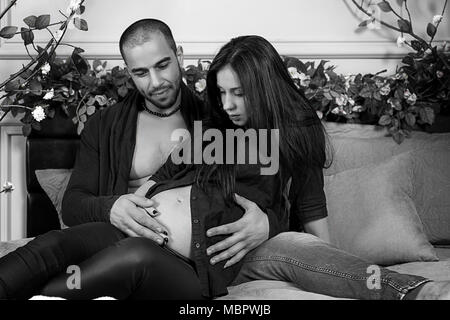 Black and white photo of muslim man and his pregnant wife, he hugging her belly while sitting on the gray cozy bed in the bedroom Stock Photo