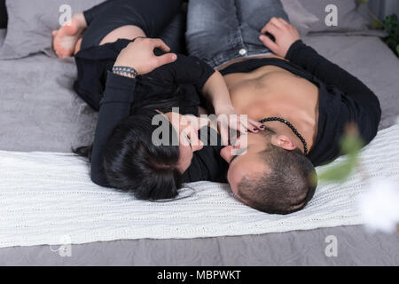 Wonderful couple of muslim man and his pregnant wife wearing black clothes, hugging while lying on the gray cozy bed in the bedroom Stock Photo