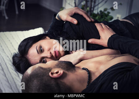 Beautiful couple of muslim man and his brunette wife wearing black clothes, hugging while lying on the gray cozy bed in the bedroom Stock Photo