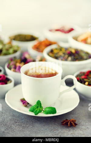 Tea concept with copy space. Different kinds of dry tea in white ceramic bowls and cup of aromatic tea on grey background. Stock Photo