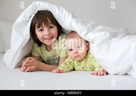 Cute little six month old baby boy and his older brother, playing under duvet at home in bed in bedroom, smiling happily Stock Photo