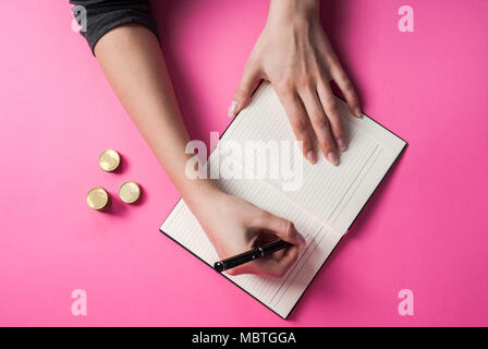 Woman hand writing with a pen in a notebook and coin on pink background Stock Photo