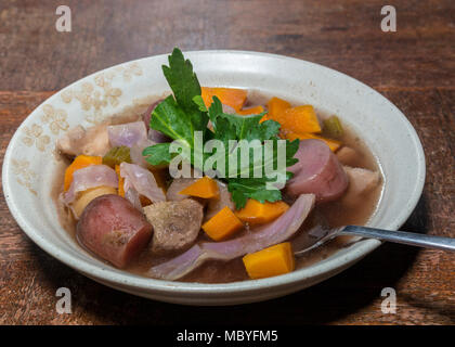Crockpot stew with potatoes, beef, red cabbage and carrots in tan bow, garnished with celery leaves, on wooden table, viewed from dinner angle . Stock Photo