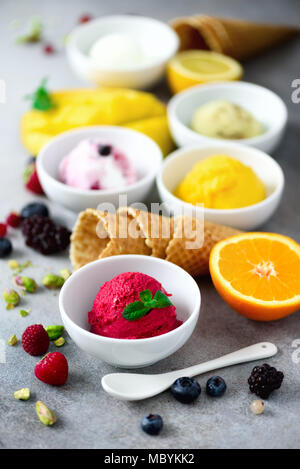 Ice cream balls in bowls, waffle cones, berries, orange, mango, pistachio on grey concrete background. Colorful collection, flat lay, summer concept Stock Photo