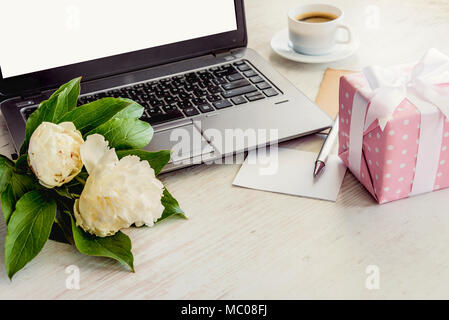 Side view of a deck with computer, bouquet of peonies flowers, cup of coffee, empty card and pink dotted gift box. White rustic wooden background. Rom Stock Photo